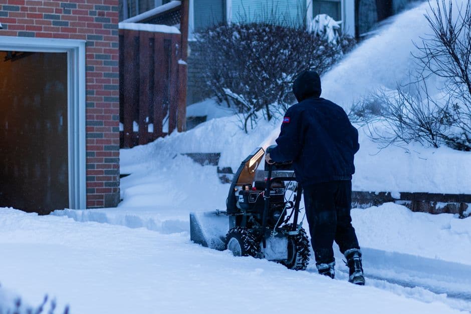A person using a snow blower to clear snow from a suburban sidewalk in winter